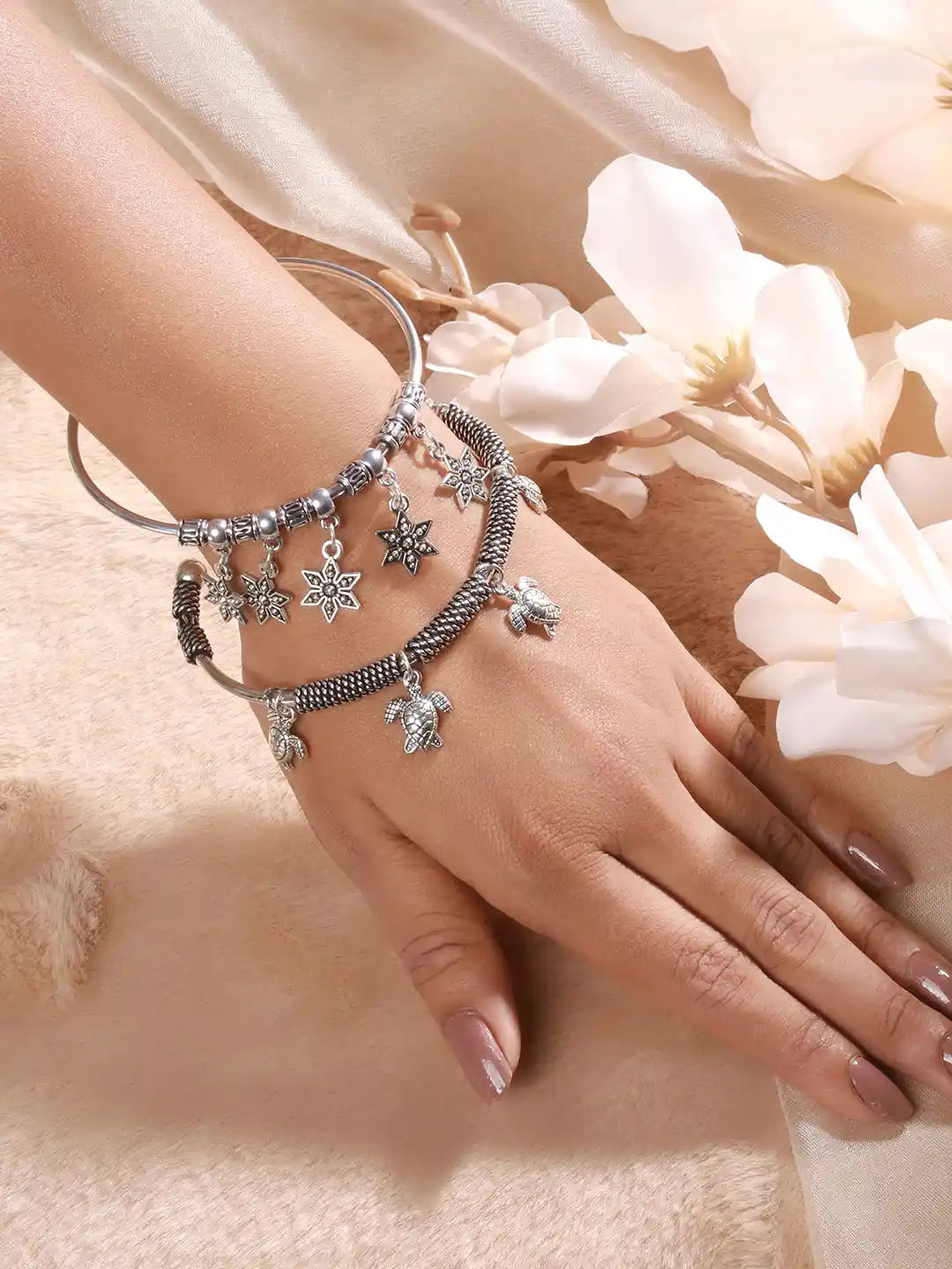 Hand wearing multiple silver bracelets with charms on a soft background with flowers