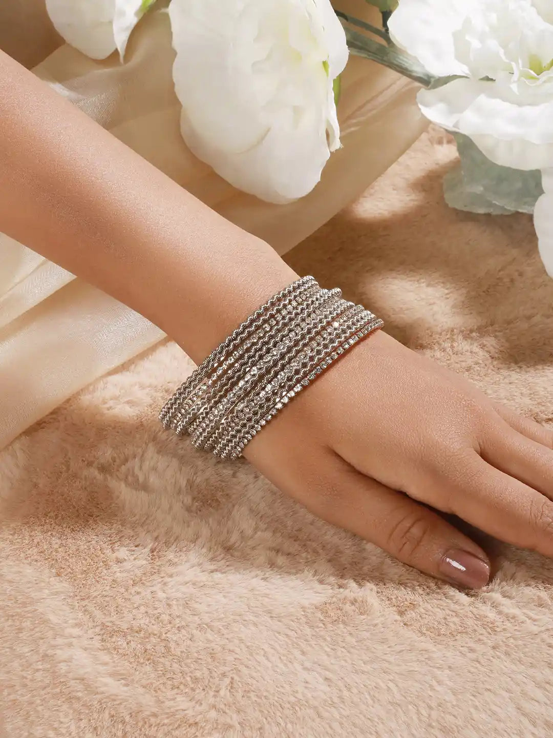 Hand wearing multiple silver bracelets on a soft surface with flowers in the background